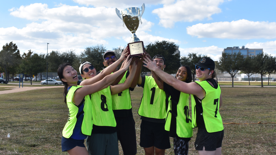 Six students stand together on a grassy field, all wearing bright yellow-green sports pinnies with black numbers, hoisting a large gold trophy with two handles and a wooden base.