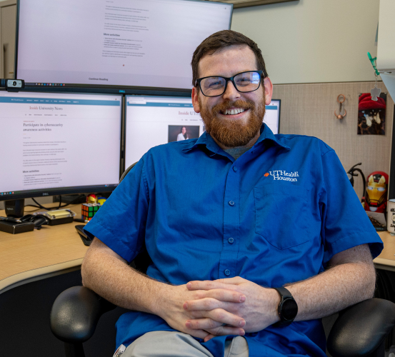 Man sits smiling at his desk in front of multiple computer monitors wearing a blue shirt.