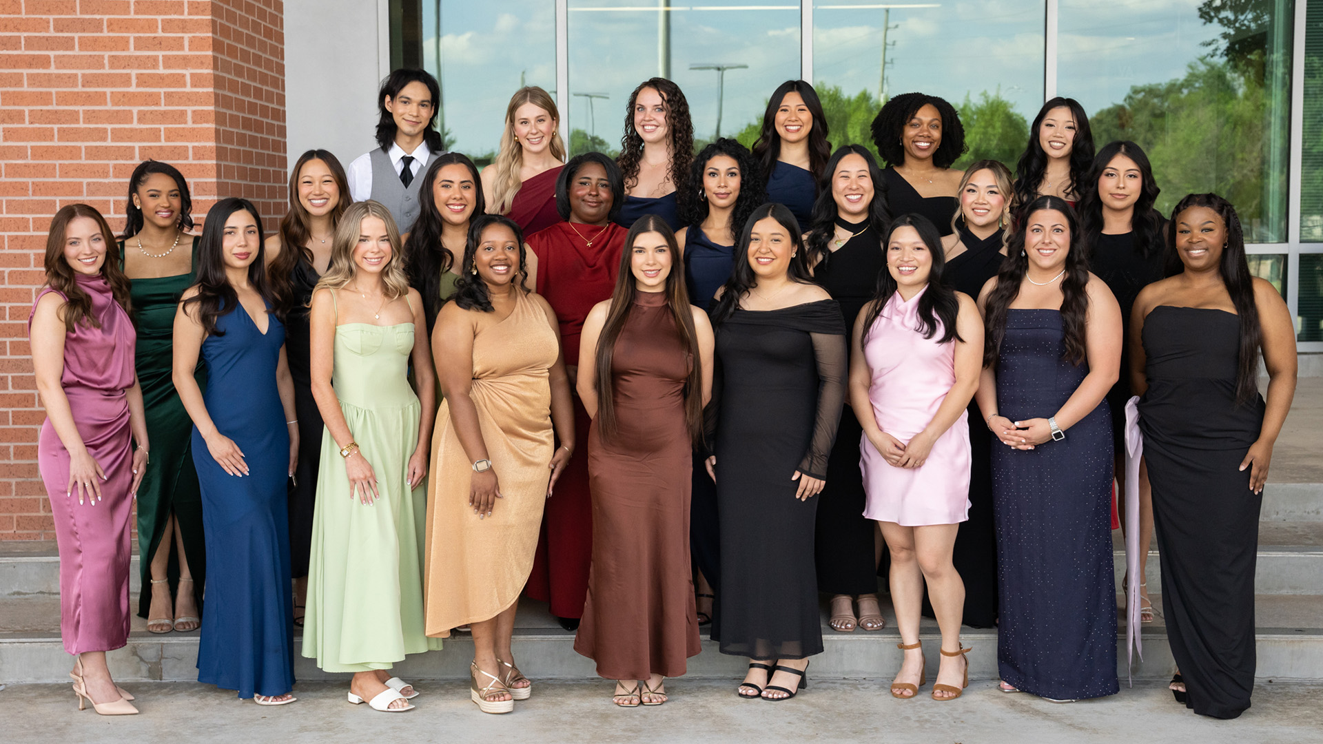 Large group portrait of individuals dressed in formal attire posing outdoors in front of a brick building and greenery.