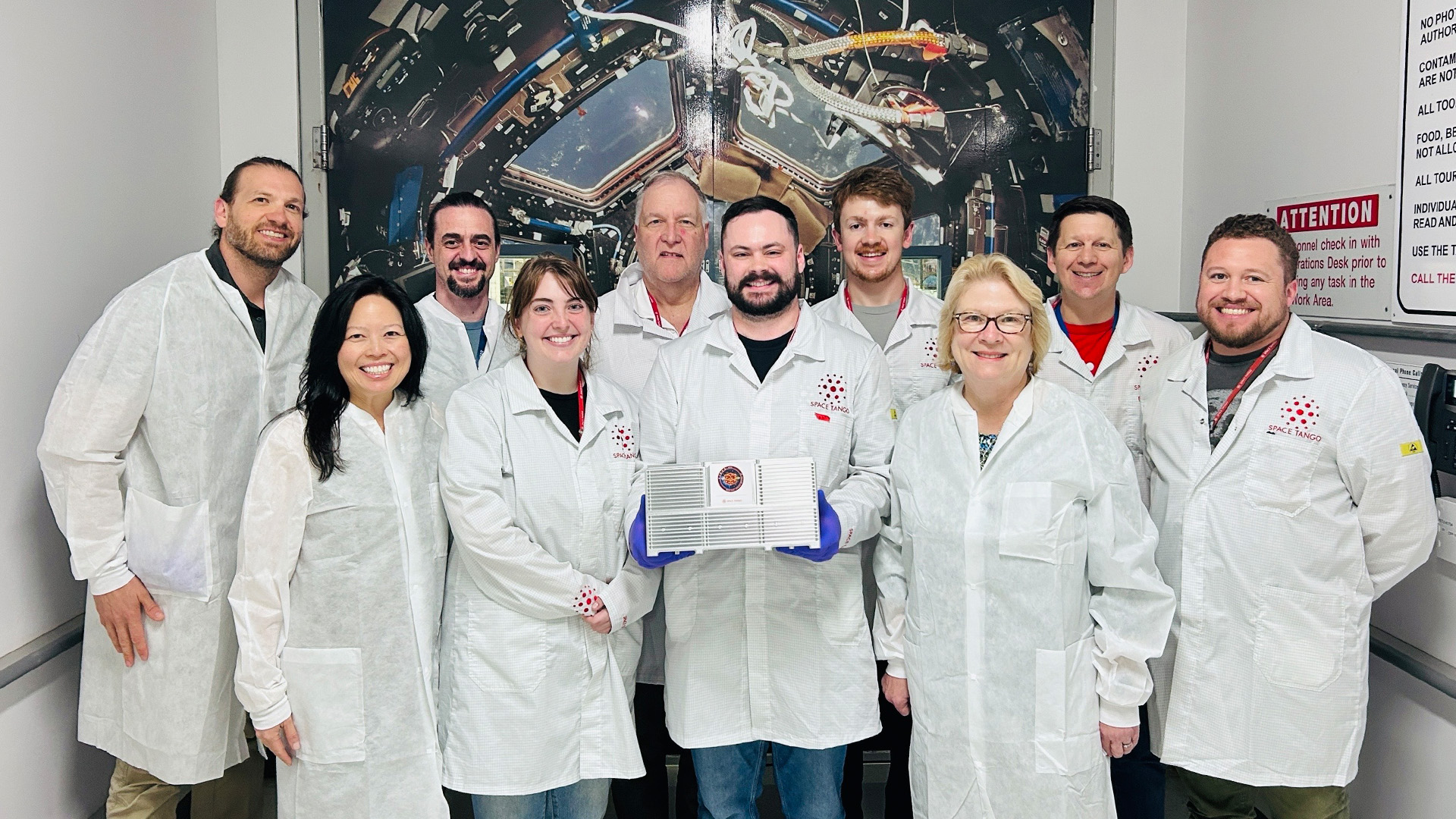 A group of ten people wearing white lab coats stand together in a laboratory or cleanroom setting. The person in the center holds a rectangular device with blue handles. Behind them is a large backdrop showing the interior of a spacecraft.