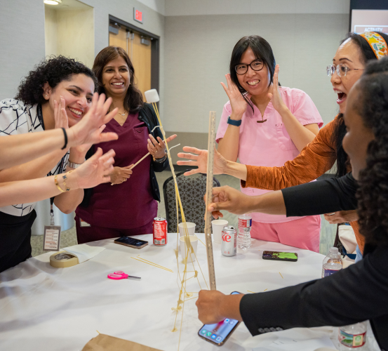 Faculty and staff cheer during the Marshmallow Challenge, a team-building activity using spaghetti, tape, and a marshmallow. One person measures their leaning tower as others laugh and react around the table.