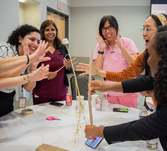 Faculty and staff cheer during the Marshmallow Challenge, a team-building activity using spaghetti, tape, and a marshmallow. One person measures their leaning tower as others laugh and react around the table.