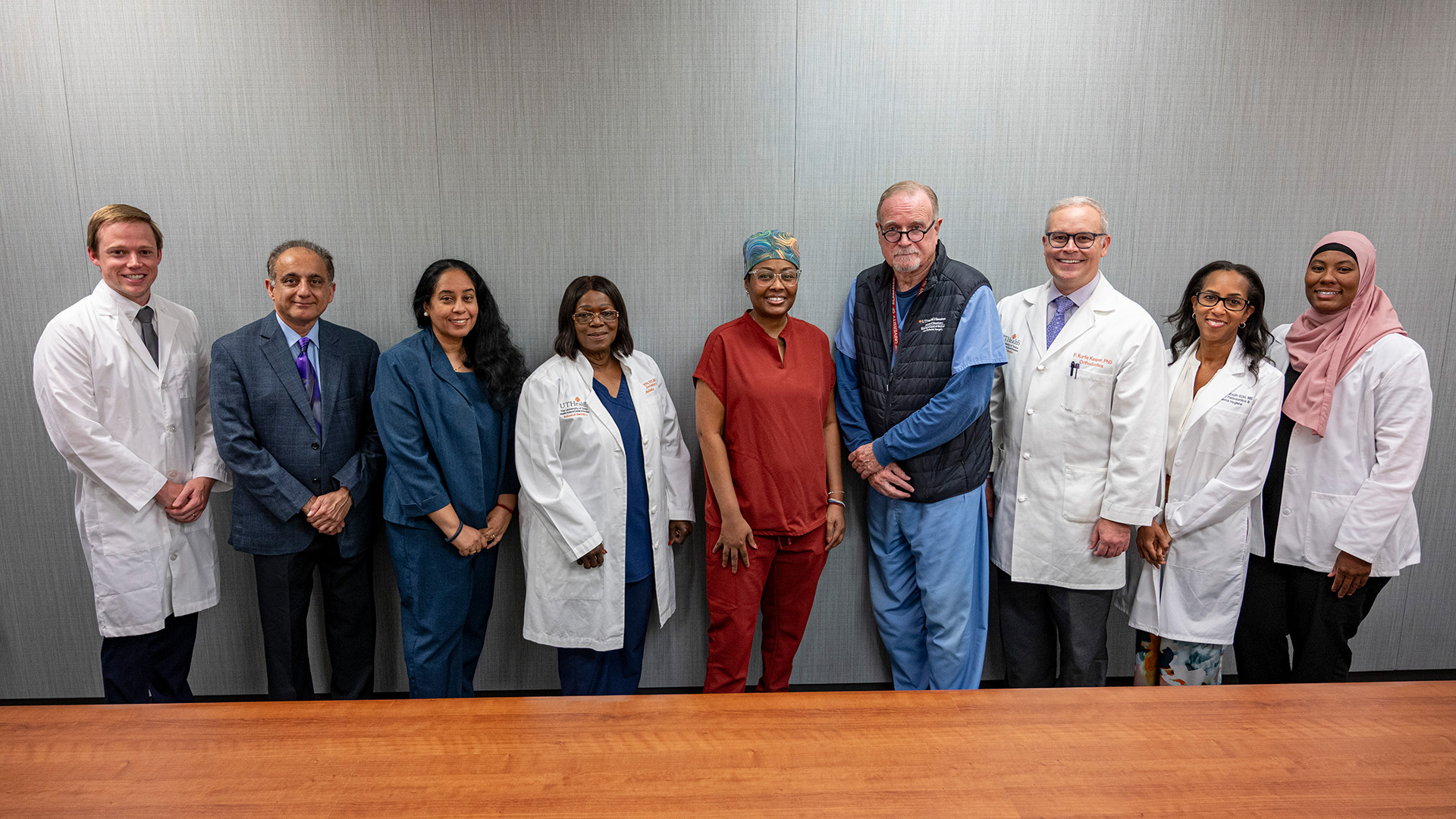 Nine individuals stand together in front of a gray wall, some wearing white lab coats, others in scrubs or professional attire, gathered behind a wooden table in a medical or clinical setting.