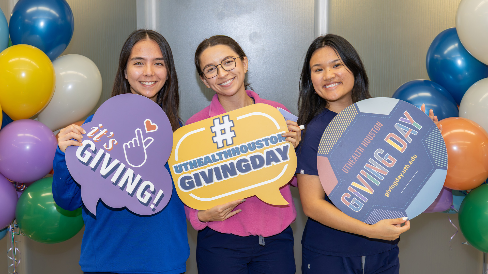 Three people stand in front of a UTHealth Houston Giving Day backdrop decorated with colorful balloons, each holding a large sign promoting the event, including “it’s GIVING!,” “#UTHealthHoustonGivingDay,” and “UTHealth Houston Giving Day.”