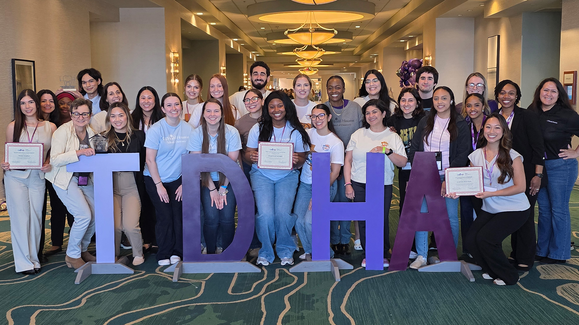 A large group of people stands together, posing behind oversized freestanding letters that spell “TDHA.” Several individuals hold certificates and awards, suggesting a recognition.