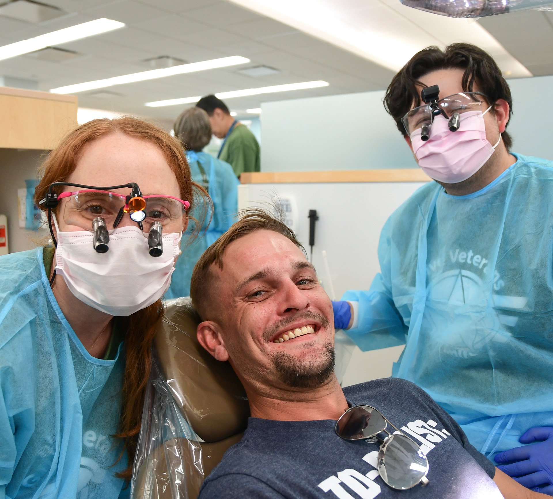 Two dental students wearing masks and protective gowns pose with a smiling patient in a dental chair during the Give Vets a Smile event at UTHealth Houston School of Dentistry.