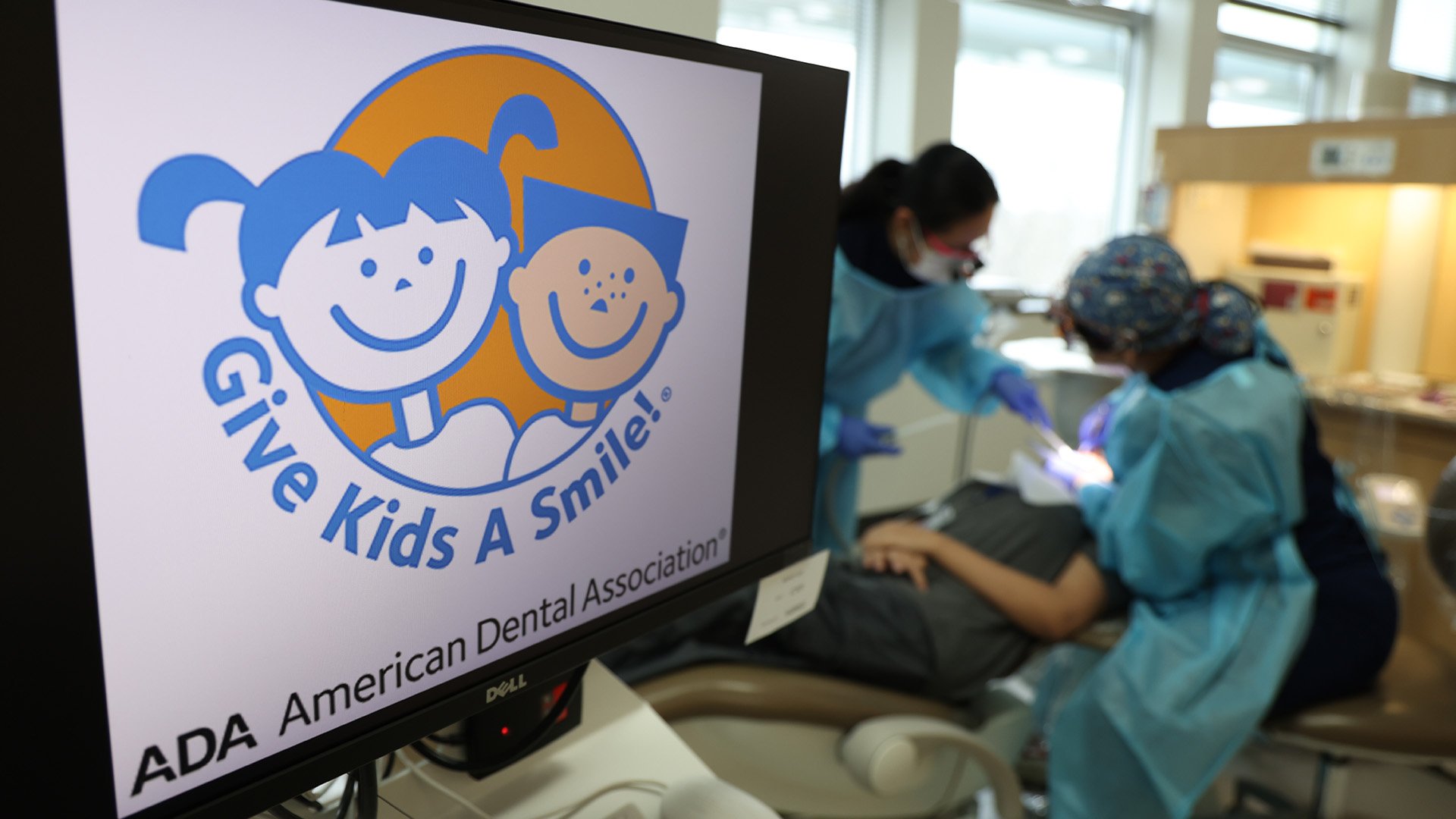 Two dental professionals attend to a patient in a dental chair. In the foreground, a computer monitor displays the Give Kids A Smile logo from the American Dental Association, featuring cartoon illustrations of a smiling boy and girl.