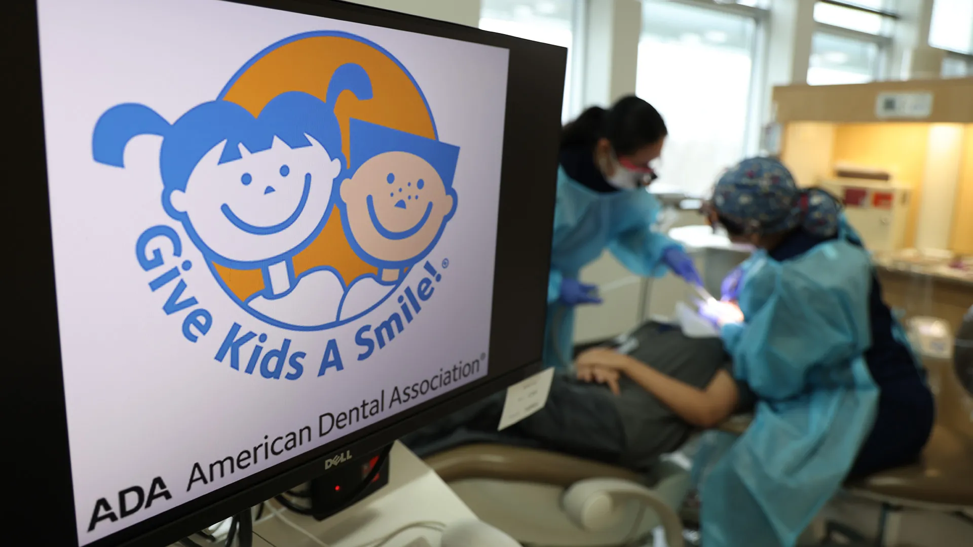 Two dental professionals attend to a patient in a dental chair. In the foreground, a computer monitor displays the Give Kids A Smile logo from the American Dental Association, featuring cartoon illustrations of a smiling boy and girl.