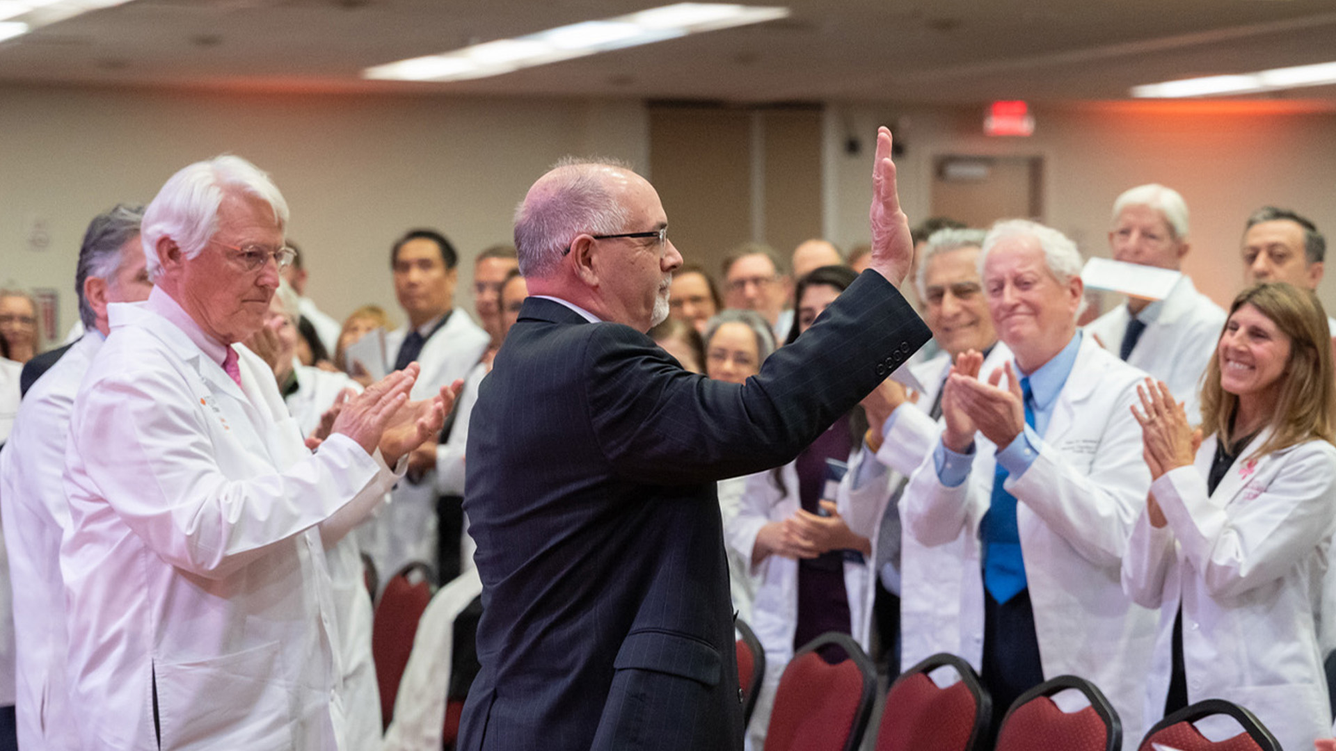 A man in a dark suit raises his hand while a group of people in white lab coats applaud around him in an auditorium setting.