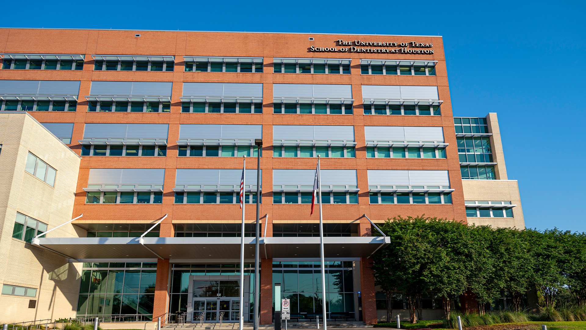 Exterior of UTHealth Houston School of Dentistry, a modern red-brick and glass structure with multiple stories, photographed on a clear day with American and Texas flags at the entrance.