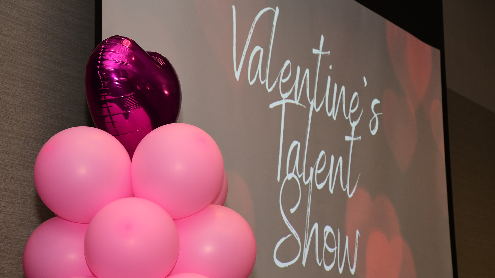 Pink balloons and a heart-shaped foil balloon beside a screen reading “Valentine’s Talent Show.”