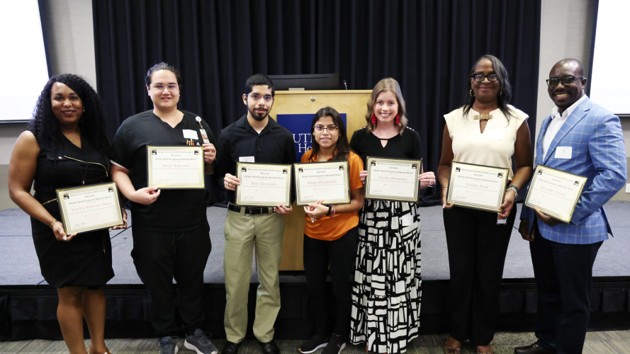 Seven people stand side by side, smiling and holding framed certificates in front of a podium and black curtain.