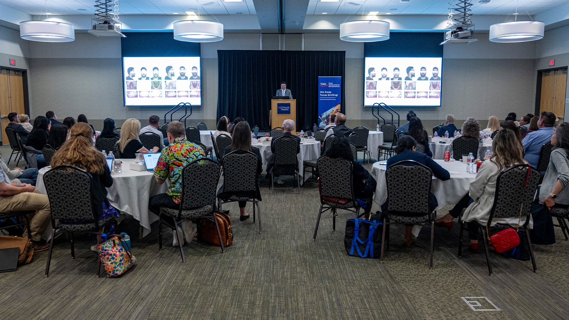 A speaker stands at the podium in a large conference room filled with round tables and seated attendees. Two large screens behind the speaker show clinical images of a patient’s face and close-up oral conditions.