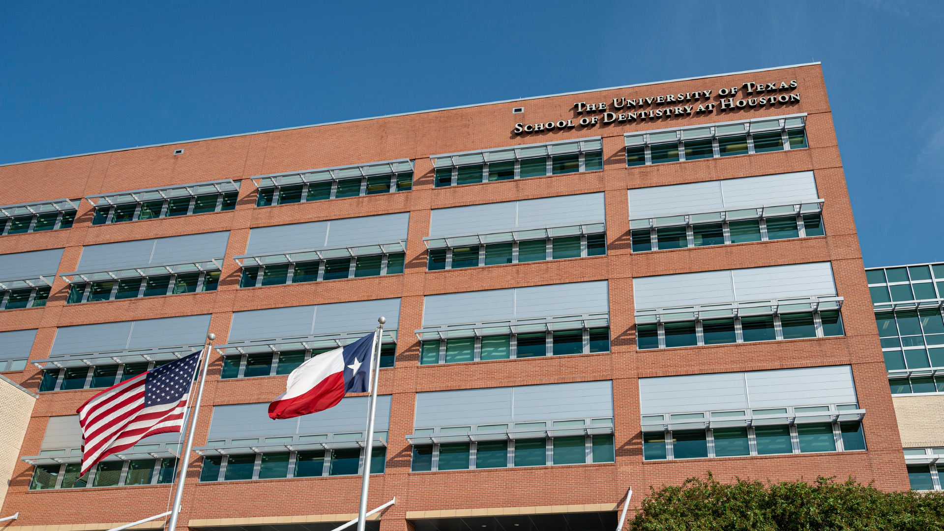 A multi-story brick building with the U.S. and Texas flags flying in front under a clear blue sky.