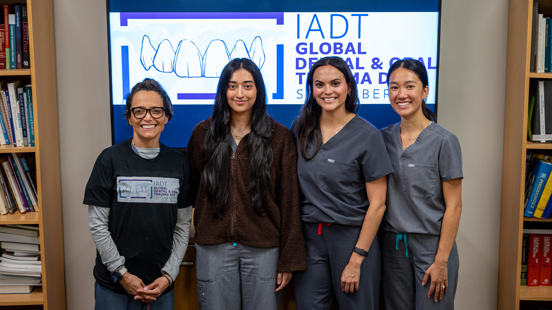Four people in scrubs and a T-shirt stand in front of an IADT Global Dental and Oral Trauma Day logo, with shelves of medical and dental textbooks behind them.