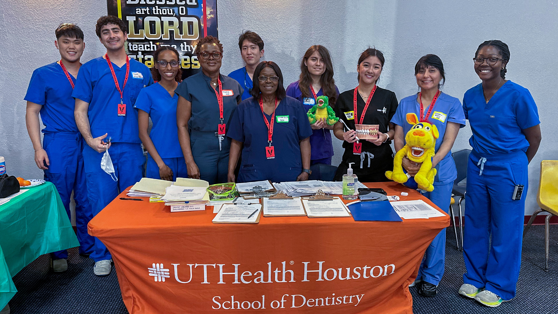 A group of 10 people in scrubs and red lanyards stand behind a UTHealth Houston School of Dentistry table displaying pamphlets, dental models, and educational materials. A wall poster is visible behind them.