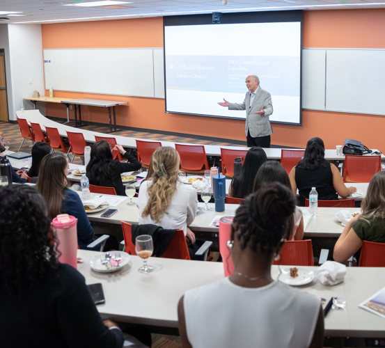 A man in a gray suit gestures while speaking at the front of a classroom during a luncheon lecture.