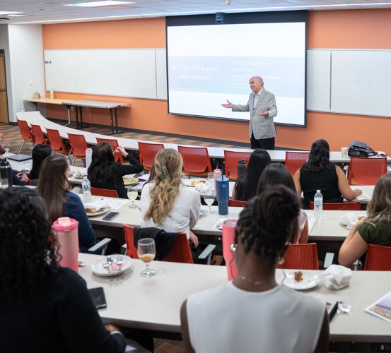 A man in a gray suit gestures while speaking at the front of a classroom during a luncheon lecture.