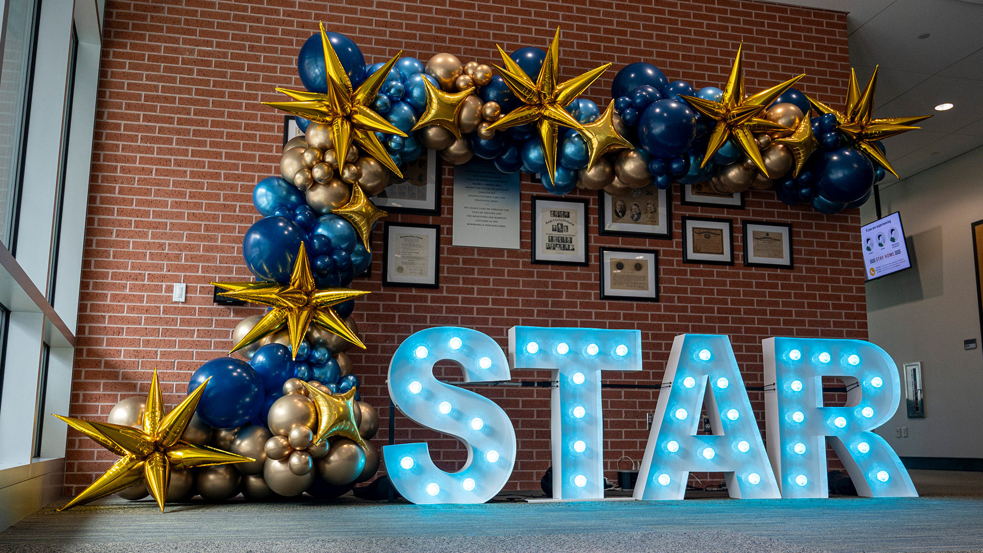 Illuminated marquee letters spelling “STAR” beneath a gold star and blue balloon display against a brick wall.