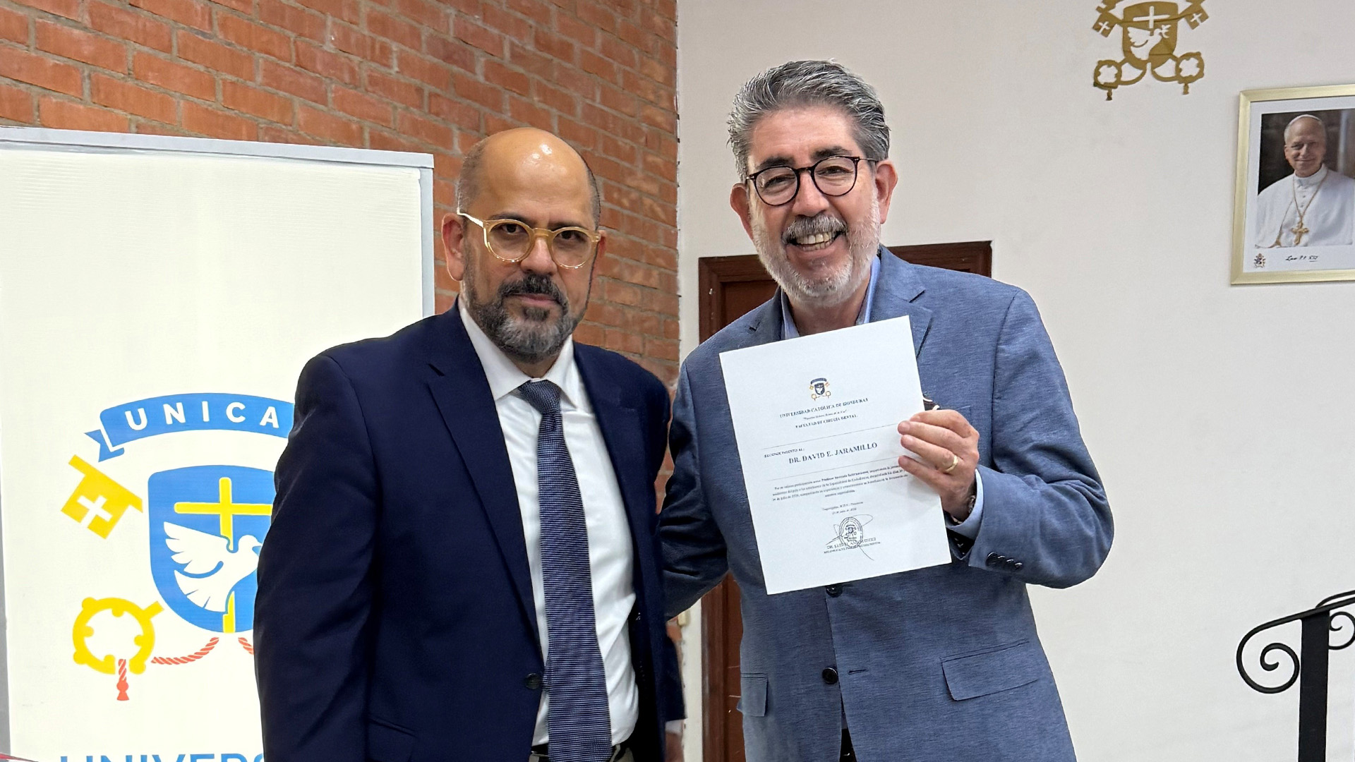 Two individuals stand indoors in front of a brick wall and a UNICA banner; one holds a certificate while both pose for a formal recognition photo.