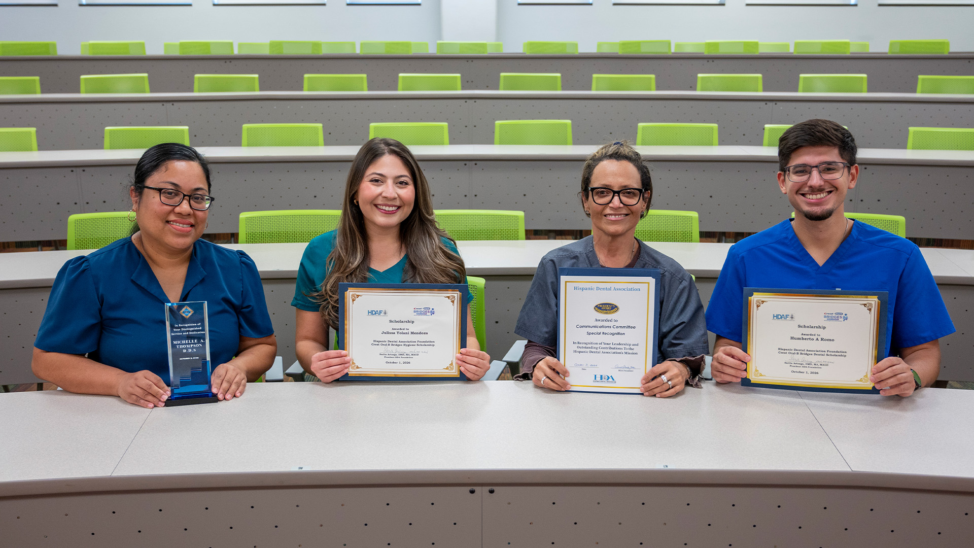 Four people seated in a lecture hall display awards and certificates during a recognition ceremony.