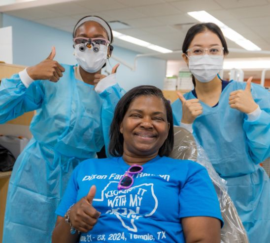 Dental providers in clinical attire and a patient seated in a dental chair pose for a photo giving thumbs up.
