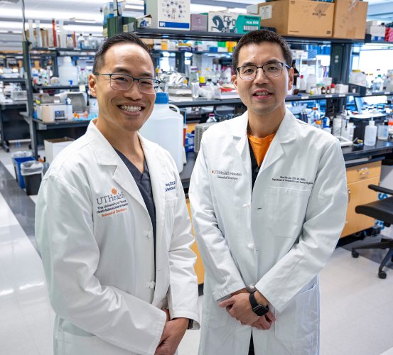 Two researchers stand in a lab wearing white coats.