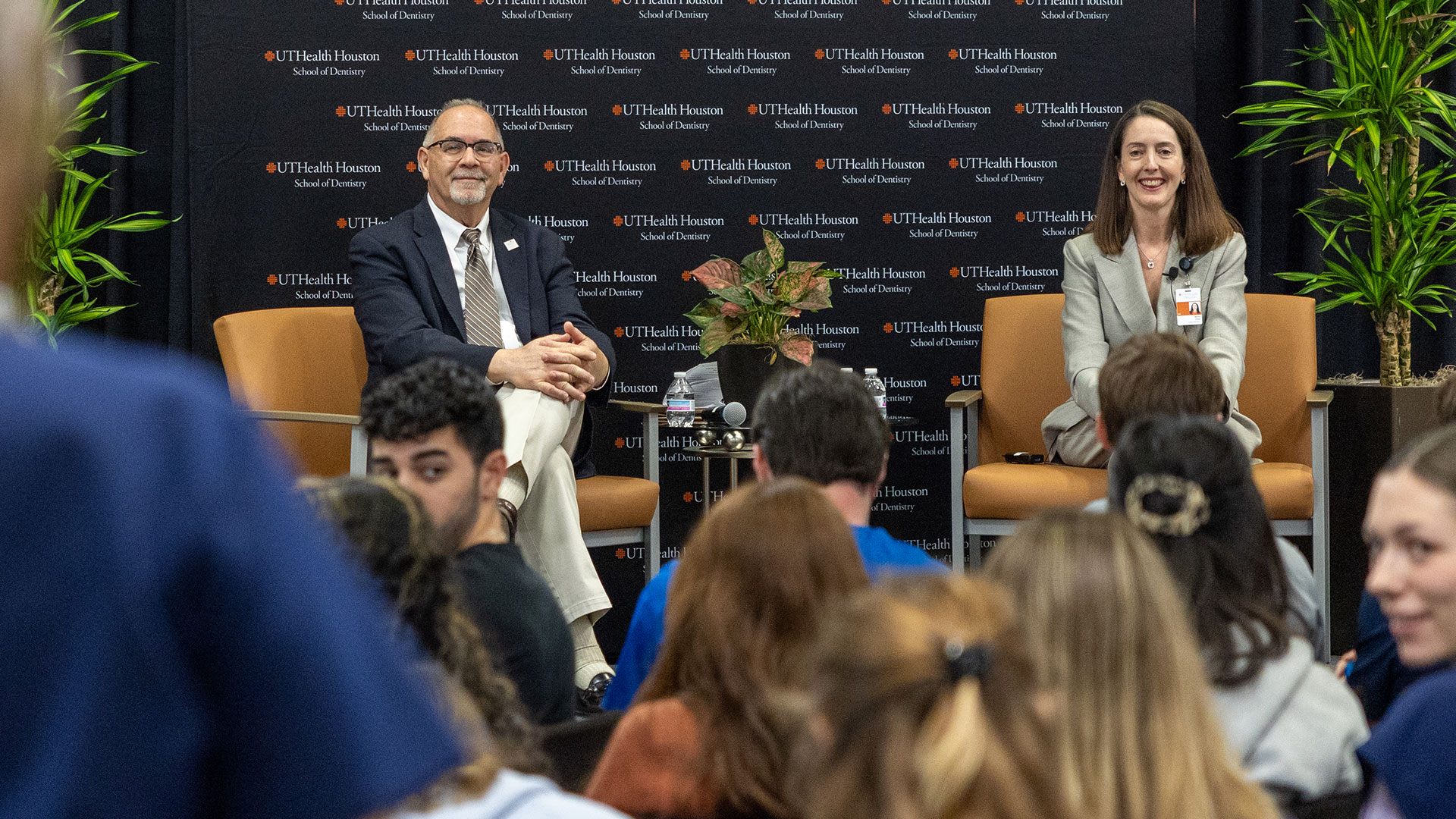 Two people in professional dress sit on stage listening to a question being asked from the audience.