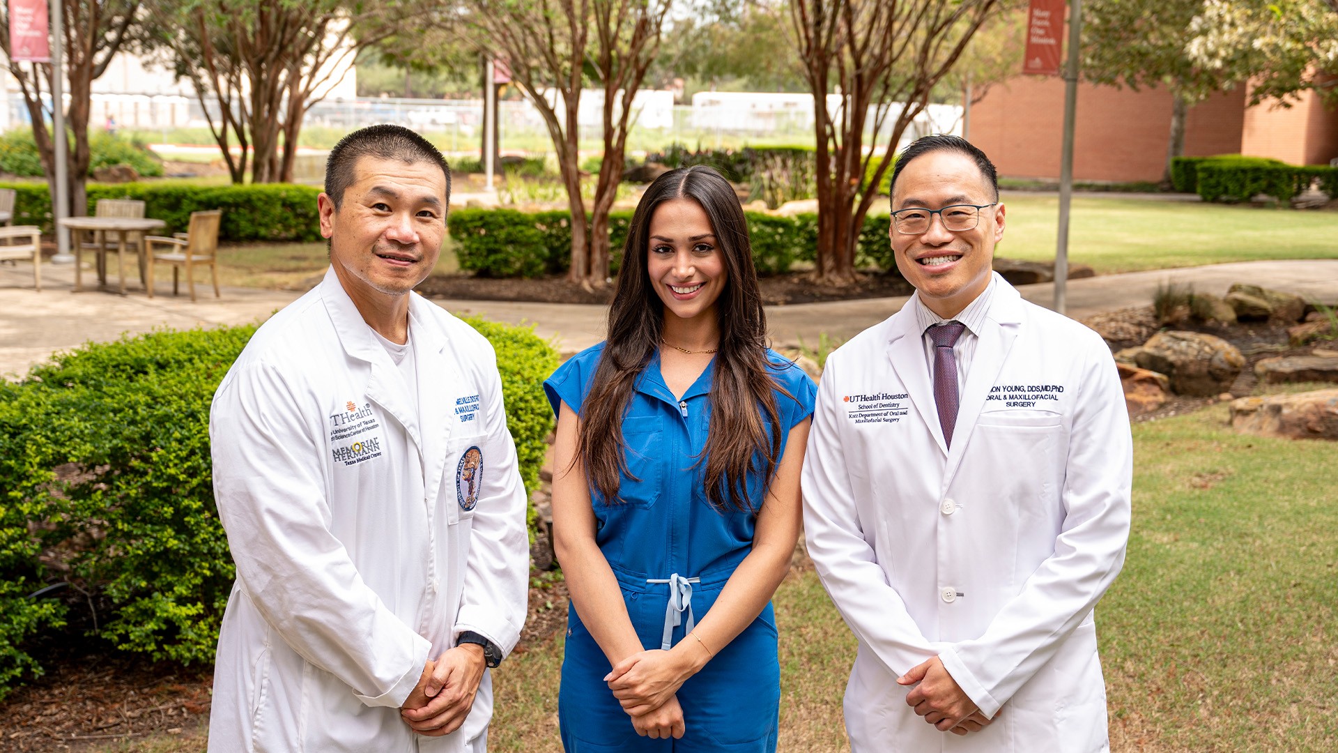 Three people pose outdoors on the UTHealth Houston School of Dentistry campus, with two individuals in white clinical coats standing on either side of a person in blue scrubs. Trees, landscaped greenery and campus walkways are visible in the background.