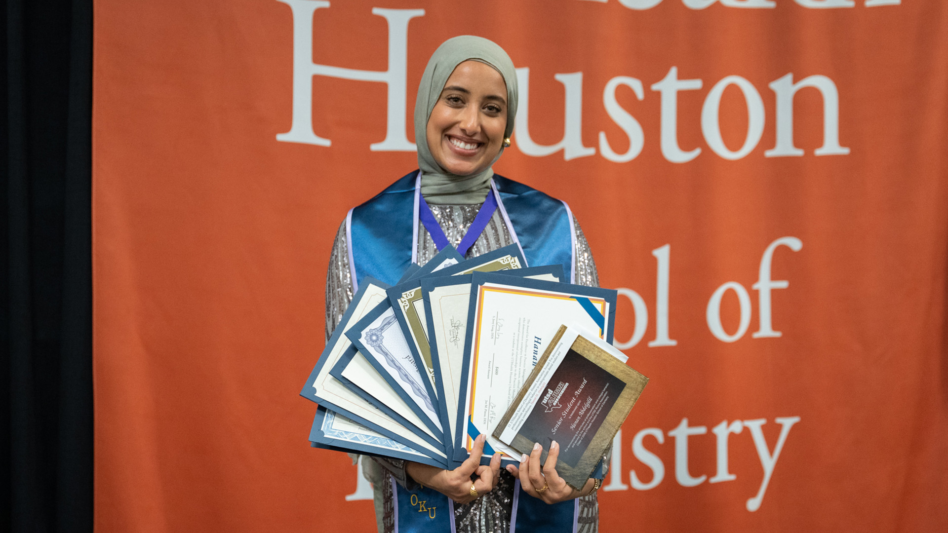 A female dental student wearing a light headscarf, silver outfit, and a blue OKU stole stands in front of an orange “Houston School of Dentistry” backdrop while holding several certificates and awards.
