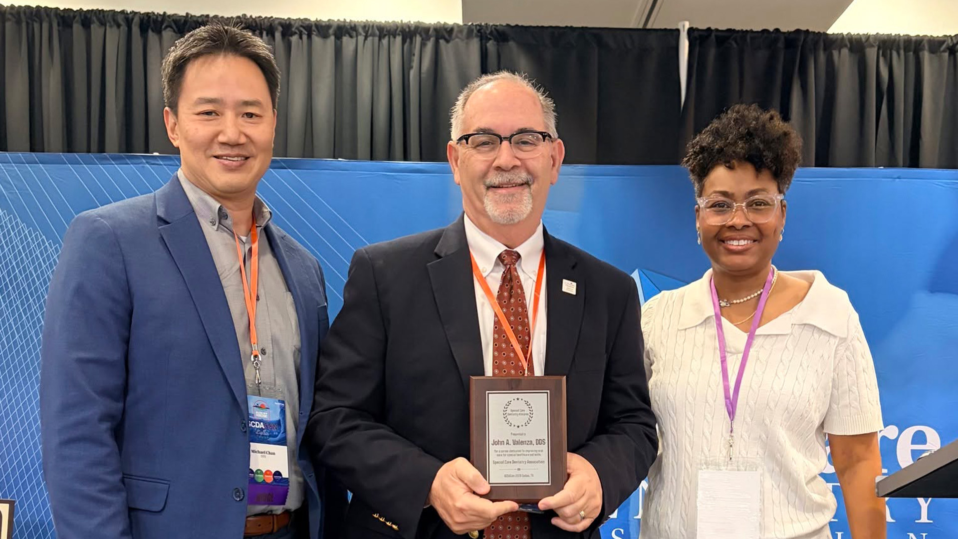 Three people stand before a blue geometric backdrop, smiling toward the camera. John A. Valenza, DDS, holds a Special Care Dentistry Association plaque recognizing his outstanding service and dedication. All wear professional attire and conference badges.