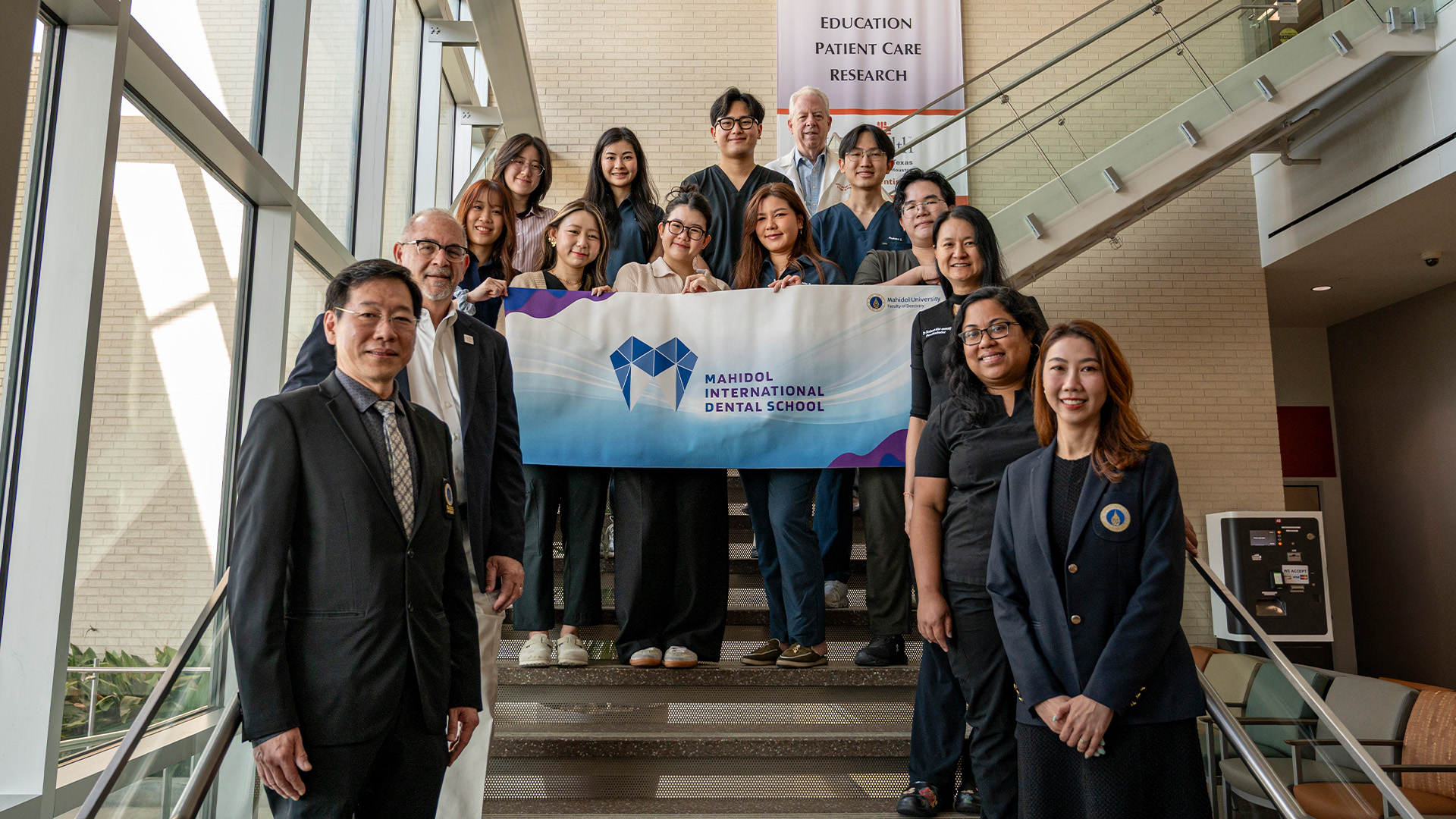 A group of people stand on an indoor staircase holding a Mahidol International Dental School banner, with large windows and a brick wall behind them.