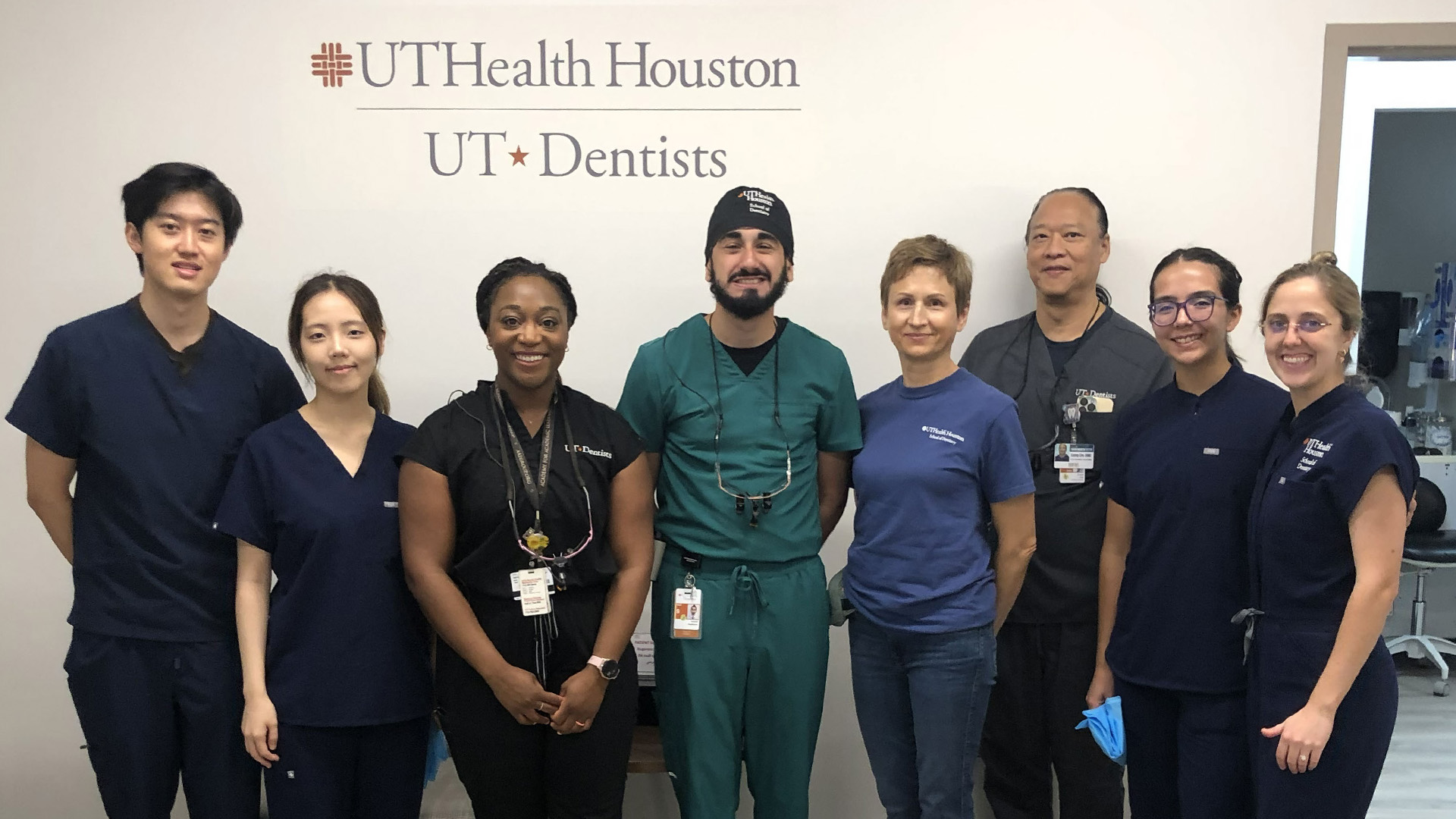 Group of eight dental professionals in clinical scrubs standing together in front of a UTHealth Houston School of Dentistry wall sign.