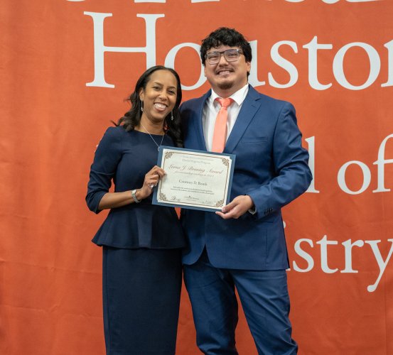 Two people pose with a certificate in front of an orange backdrop.