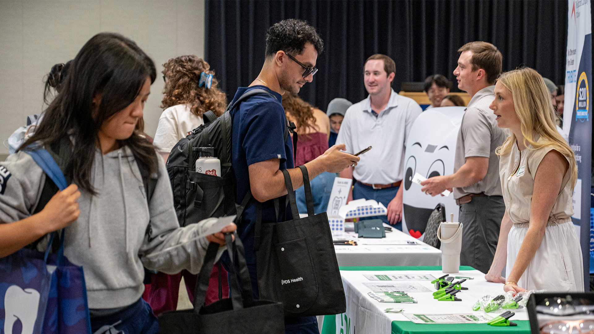 Visitors gather around an information table at an indoor event, speaking with representatives and reviewing materials and promotional items.