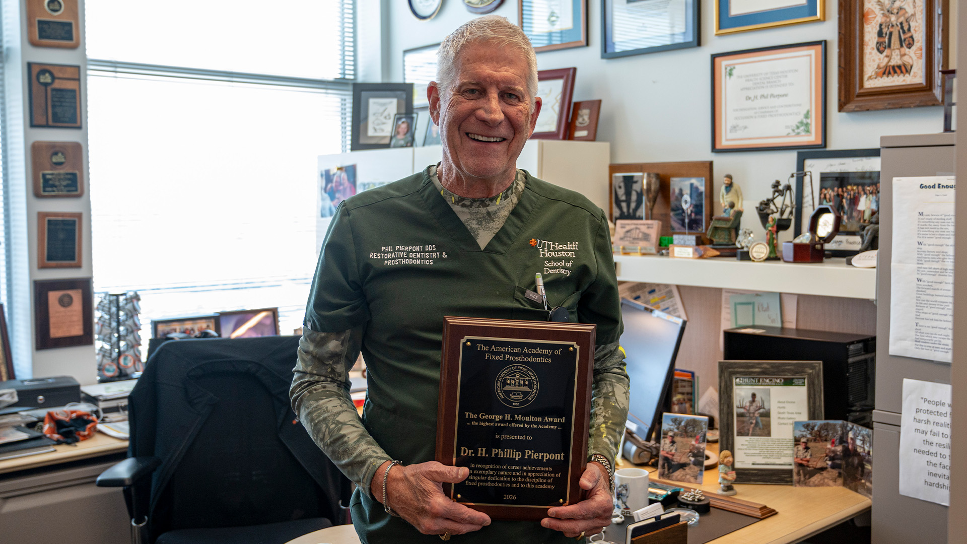 A man wearing a green UTHealth Houston School of Dentistry scrubs in an office lined with awards, certificates, and photos, holding a plaque recognizing him.