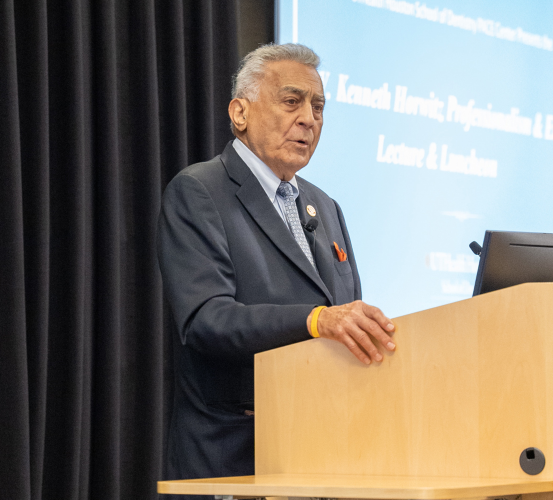 An older man in a dark suit, light blue tie, and orange pocket square speaking from a wooden podium in front of a projector screen.