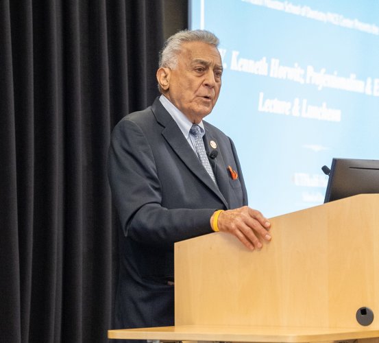 An older man in a dark suit, light blue tie, and orange pocket square speaking from a wooden podium in front of a projector screen.