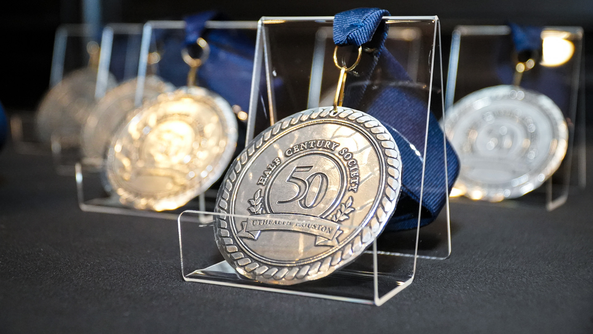 Silver commemorative medallions with engraved designs hang from blue ribbons and are displayed in clear stands on a dark tabletop.
