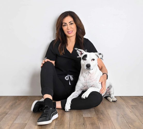 Person in black scrubs sitting cross-legged on a wooden floor beside a white dog with black spots, both facing forward against a plain white wall. The dog leans gently against the person, appearing calm and relaxed.