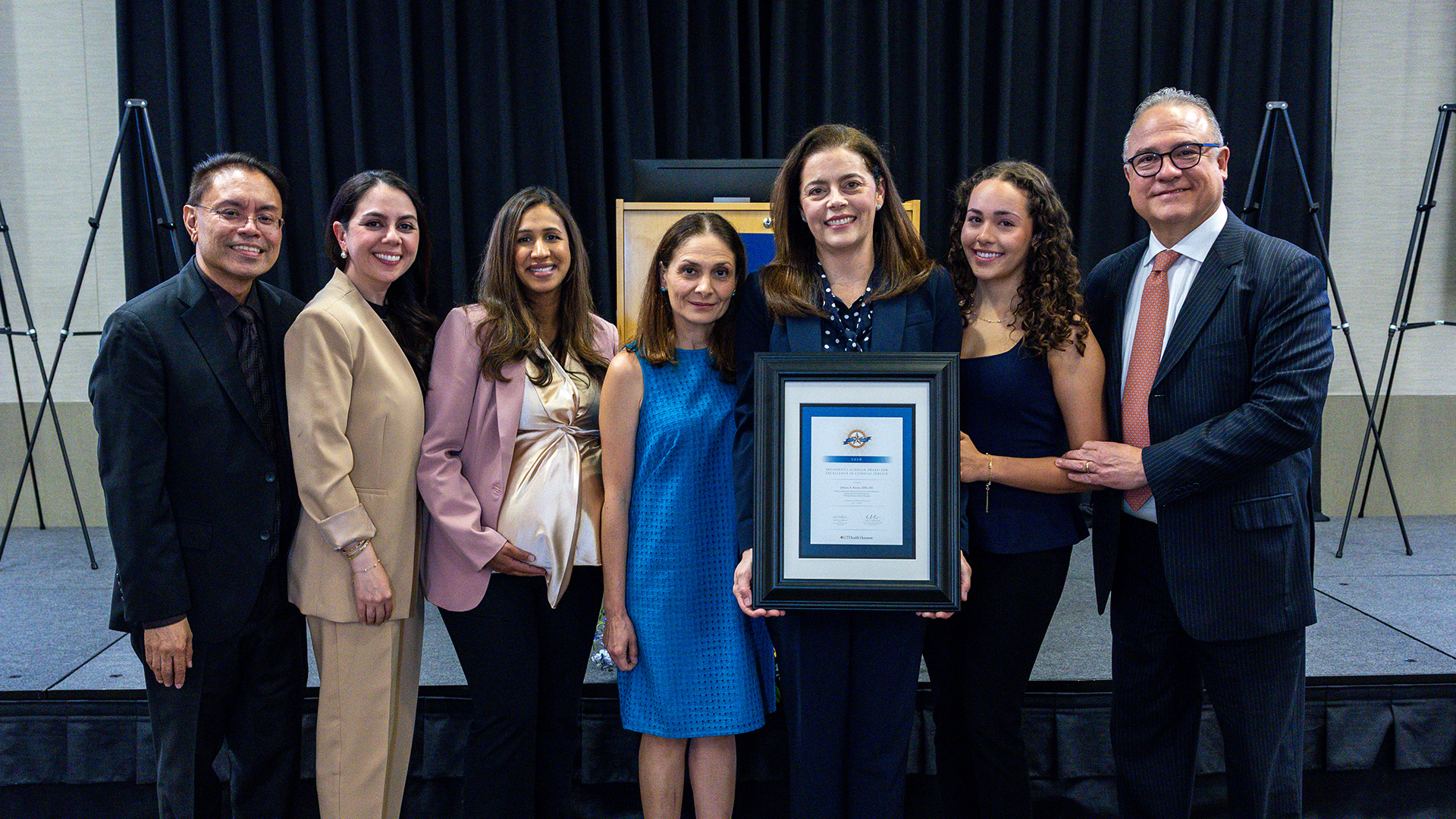 Seven people stand together on a stage in front of a black curtain. One person holds a framed certificate or award while the group poses for the photo.