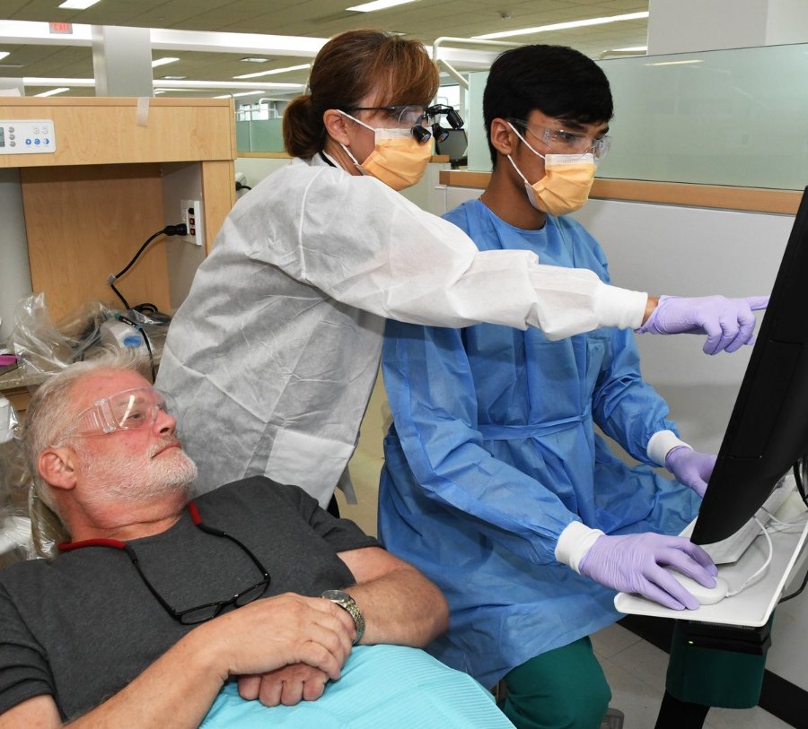 In a clinic bay, a dental hygiene instructor points to a computer screen as a dental hygiene student looks on. both are in masks, scrubs, and gloves.
