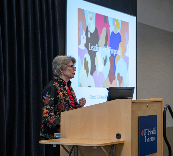 A woman wearing a black jacket with colorful floral embroidery and glasses speaks at a podium next to a projector screen during the Women in Dentistry: Leading with Purpose luncheon at UTHealth Houston School of Dentistry.