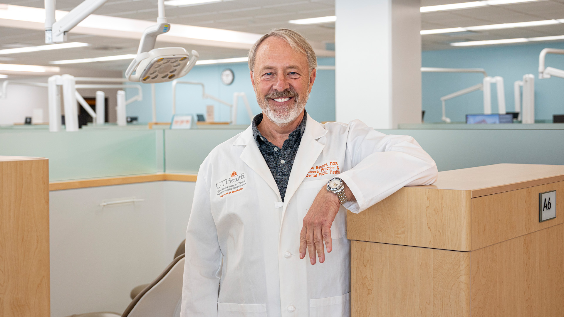 Person in a white lab coat standing in a bright, modern dental clinic, leaning on a partition with treatment areas and equipment visible in the background.