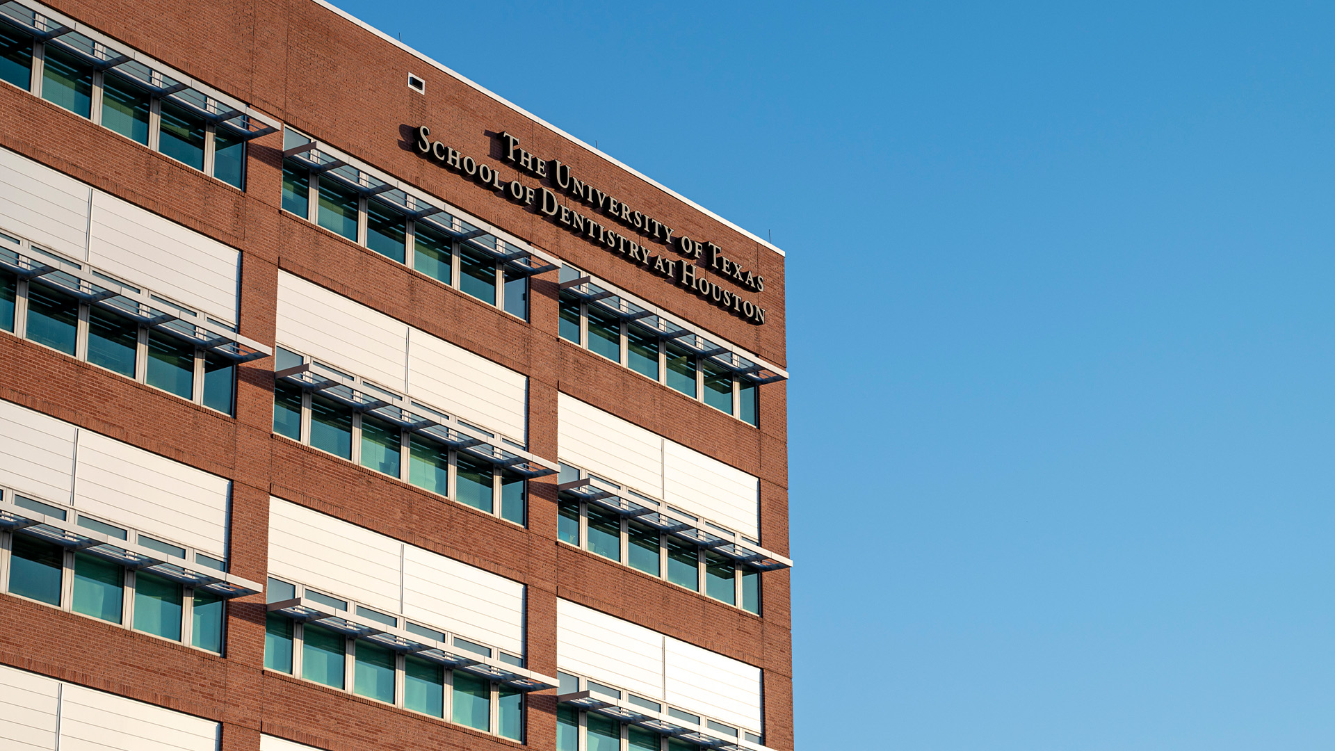Exterior of UTHealth Houston School of Dentistry. Lettering on the building says The University of Texas School of Dentistry at Houston. A blue sky is seen in the background.