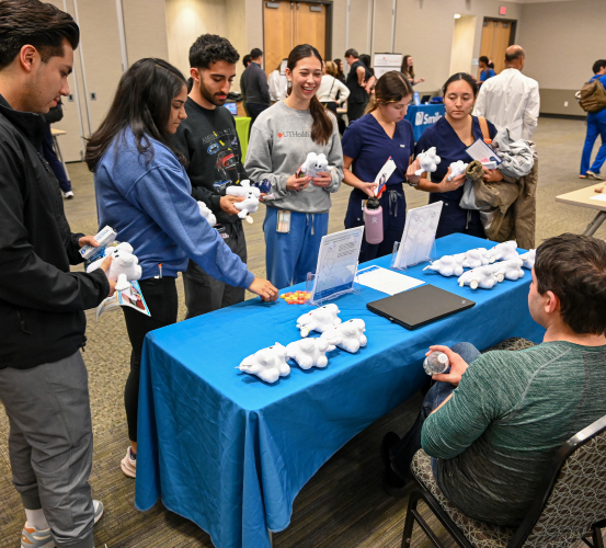 A group of students gathers at a blue-covered vendor table during a campus career fair holding small plush tooth toys and pamphlets while engaging with a seated representative.