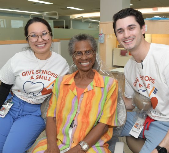 Two dental students pose for a group photo with a senior patient in a clinical setting.