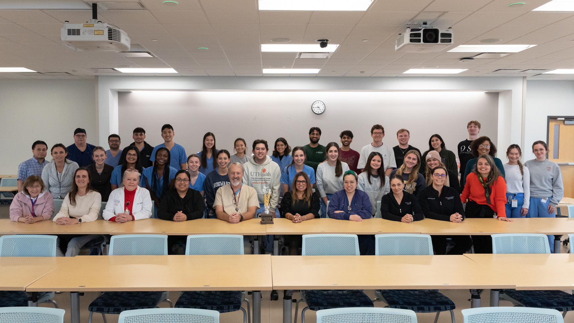 A large group of people stands together in a classroom, posing for a group photo behind several long tables.