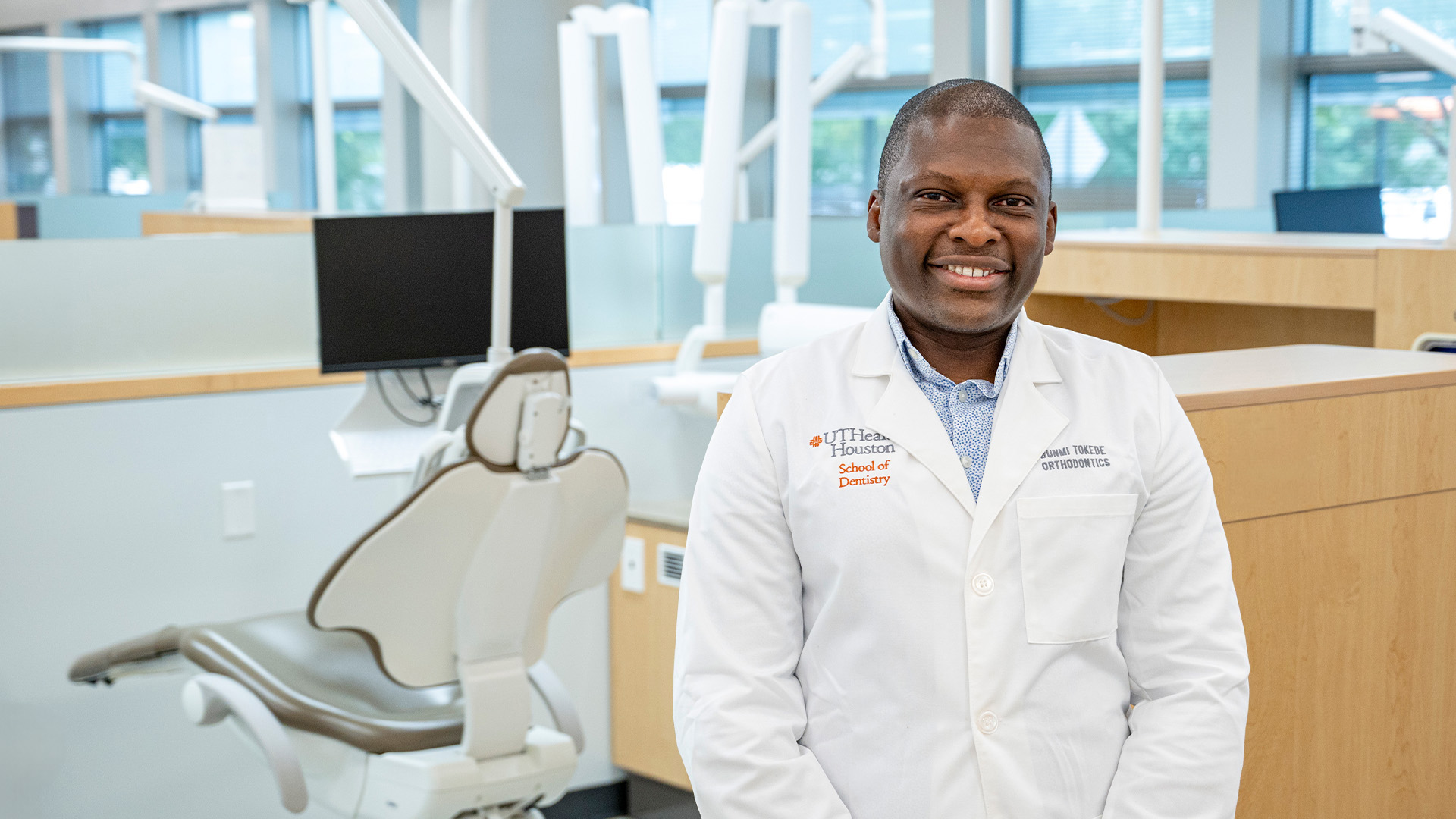 A person wearing a white lab coat stands in a bright, modern dental clinic. Dental chairs, computer monitors, and equipment fill the clean, well-lit space, with large windows in the background.