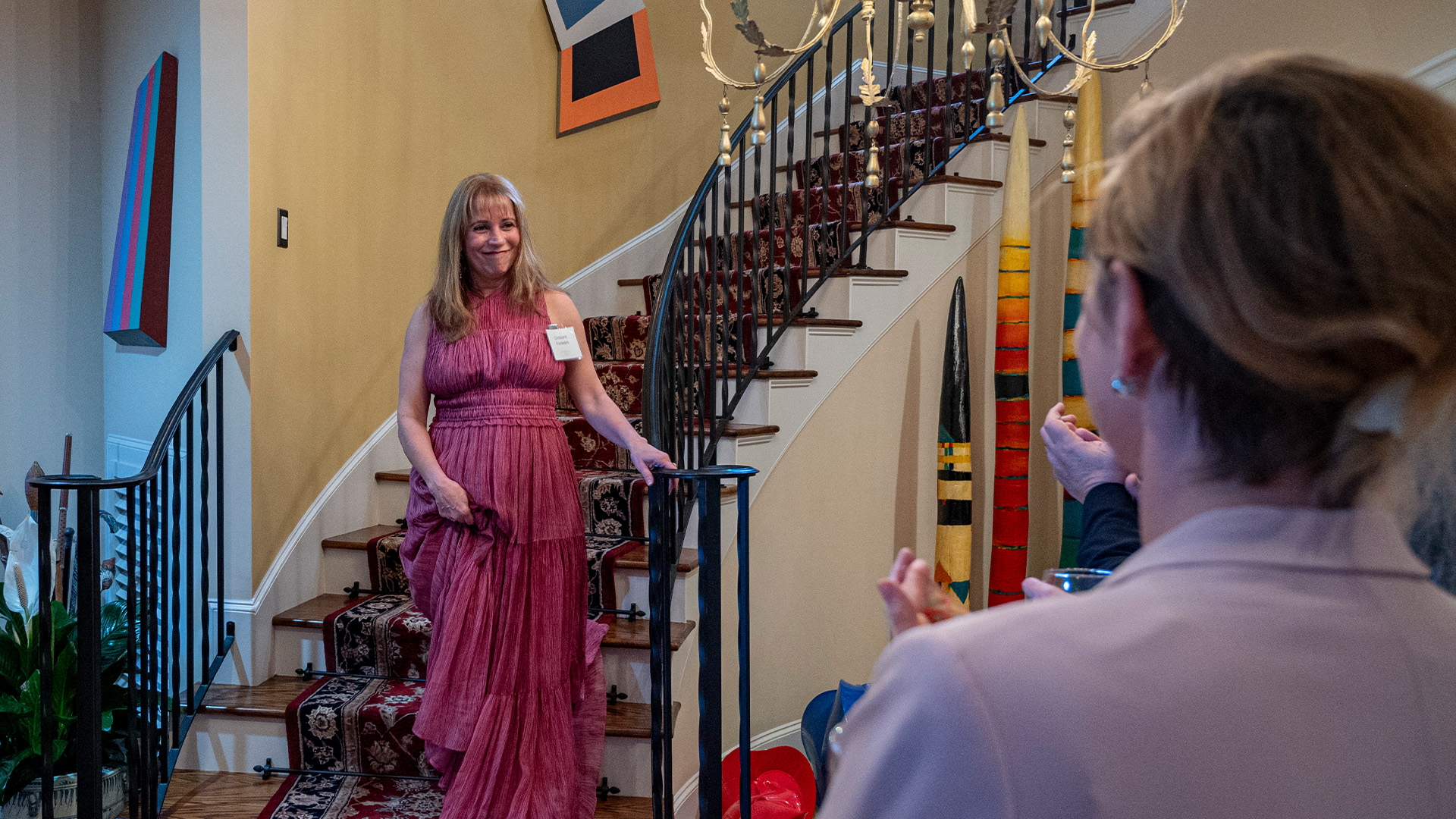 A woman in a long pleated dress walks down a curved staircase, holding part of the dress and the railing. A person in the foreground claps or gestures toward her. Colorful abstract art, tall striped sculptures, and a gold chandelier fill the space.